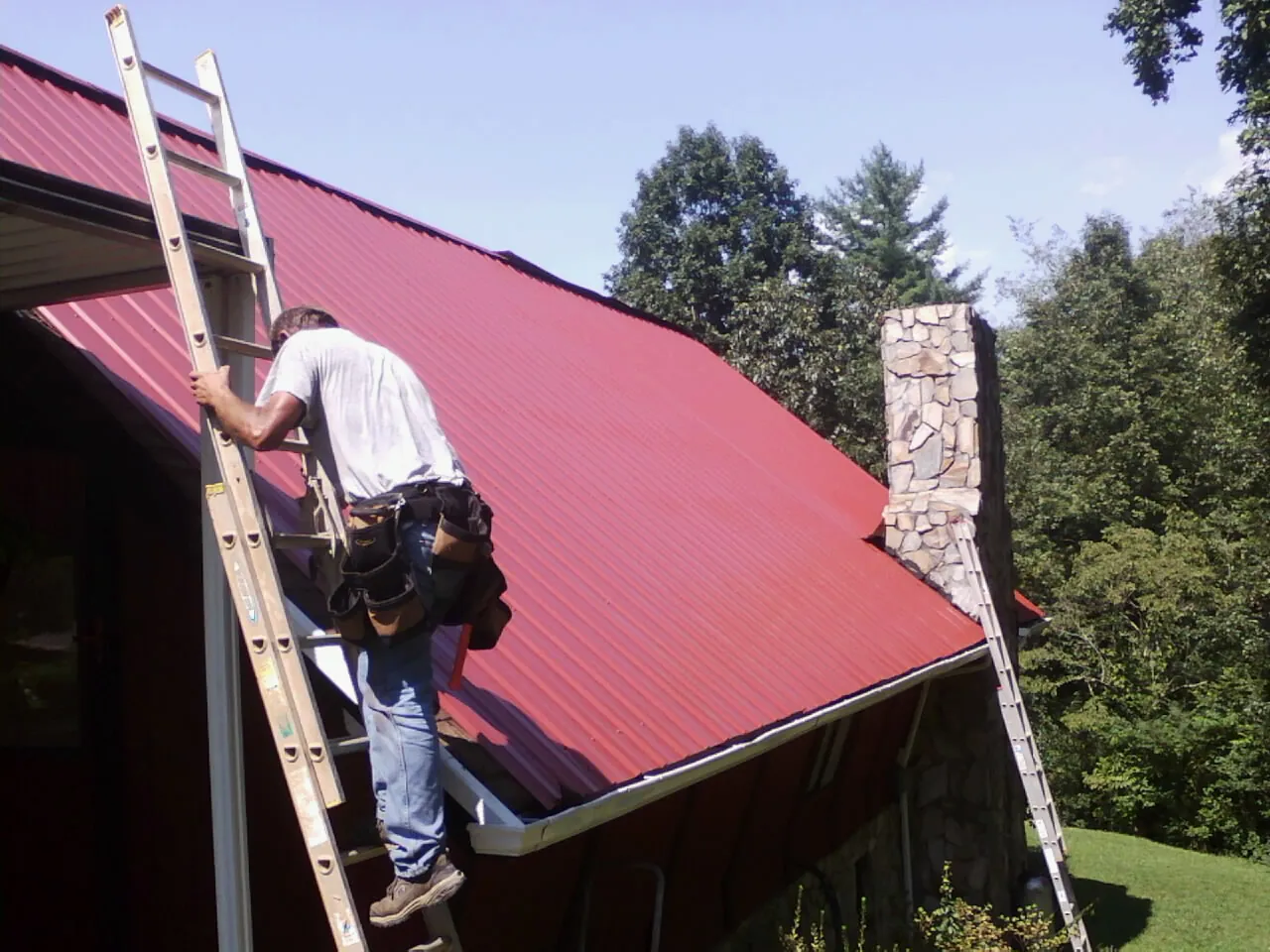 Roofer on a ladder beside a red metal roof and stone chimney — Soffit & Fascia Repair by Roofing Brothers in Housatonic
